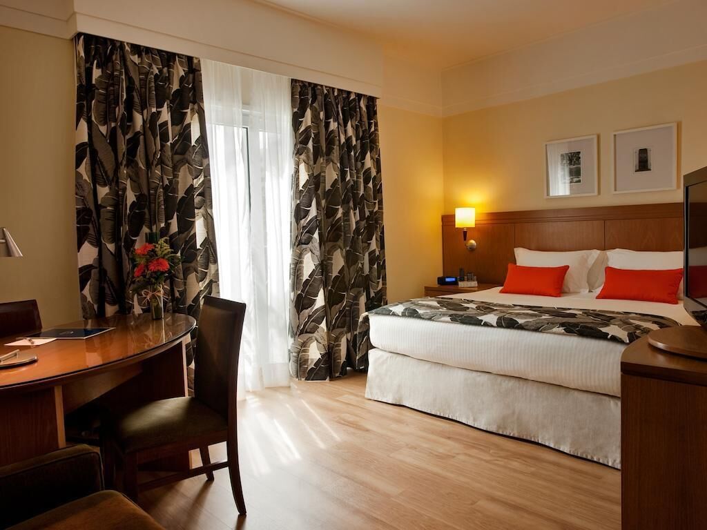 Ett hotellrum med en säng, ett skrivbord, en stol, ett nattduksbord och en lampa.