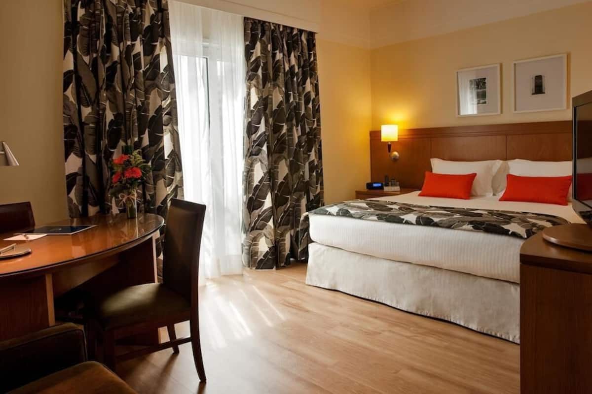 A hotel room with a bed, a desk, a chair, a nightstand, and a lamp.