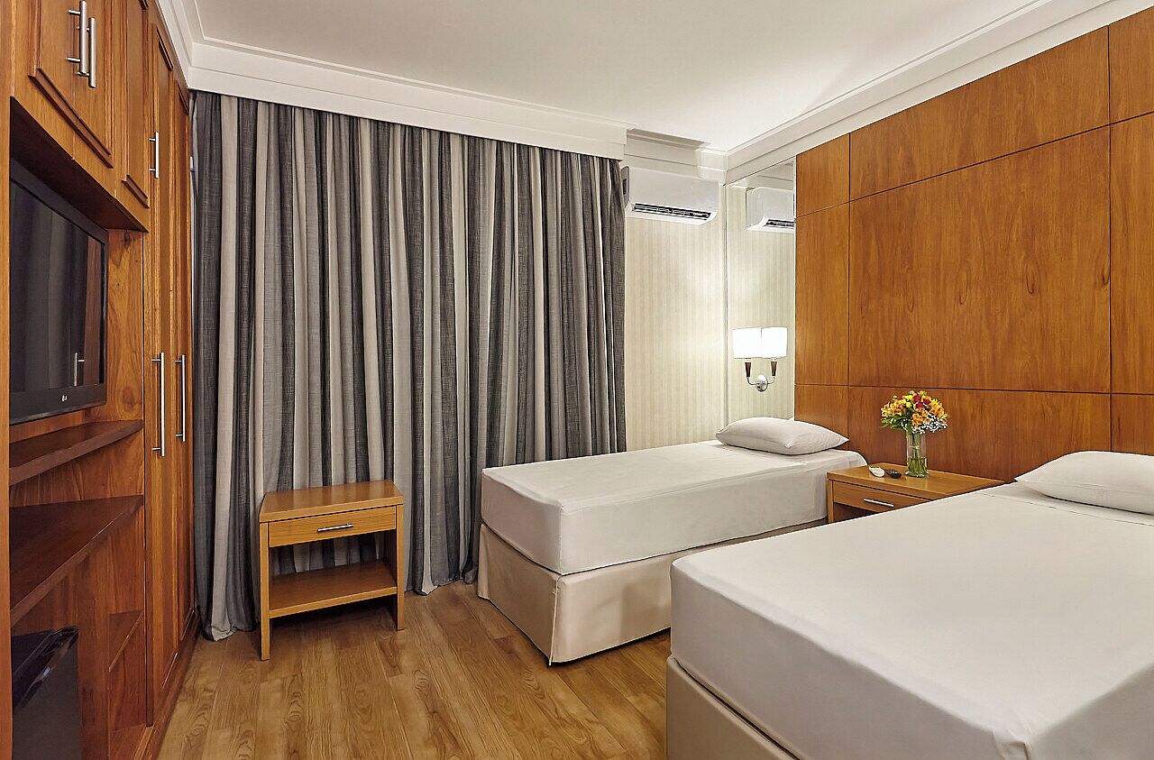 A hotel room with two beds, a wooden wardrobe, a television, a bedside table with a lamp, and a window with curtains.