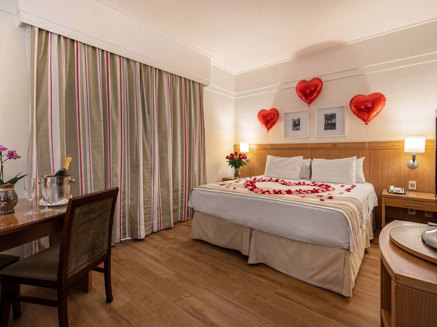Habitación de hotel con cama, escritorio, silla, mesa de comedor y un ventanal con cortinas.