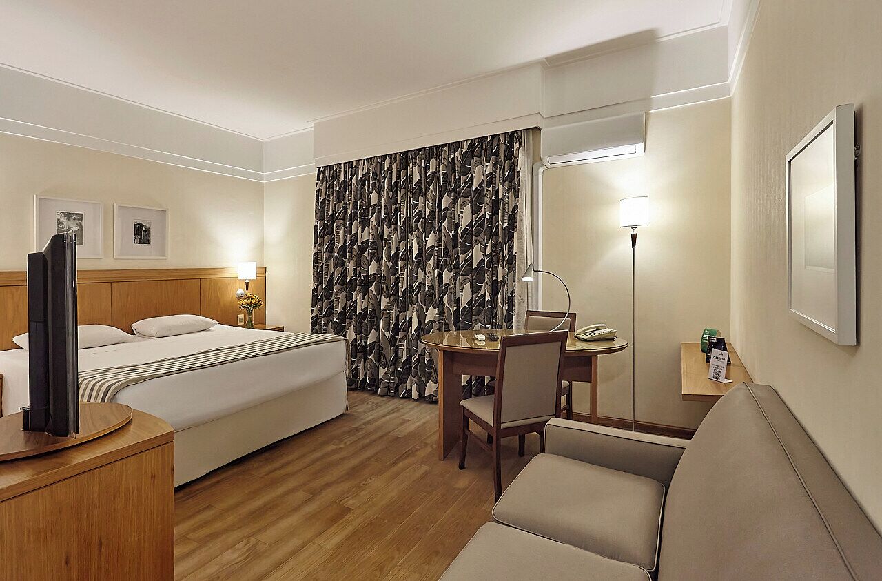 Quarto de hotel com cama, escrivaninha, cadeira, televisão, abajur e espelho.
