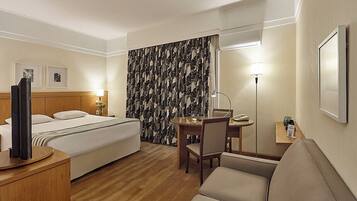Quarto de hotel com cama, escrivaninha, cadeira, televisão, abajur e espelho.