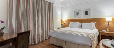 A hotel room with a wooden bed, a desk, a chair, a nightstand, and a lamp.