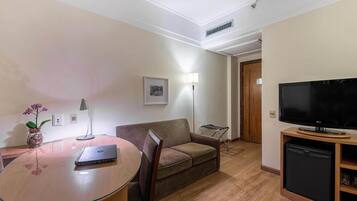 A hotel room with a wooden dining table, a laptop, a sofa, a TV, and a mini-fridge.