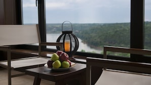 Una habitación con una mesa que sostiene un plato de manzanas, una lámpara de mimbre y vistas a un río y un bosque a través de ventanas amplias.