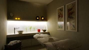 A small, dimly lit room with a single bed, a small table with a bowl and a red candle, and a window with plants.