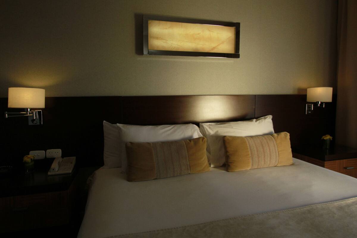 A hotel room with a large bed, two bedside lamps, a framed artwork above the bed, and a nightstand with a phone and a small vase.