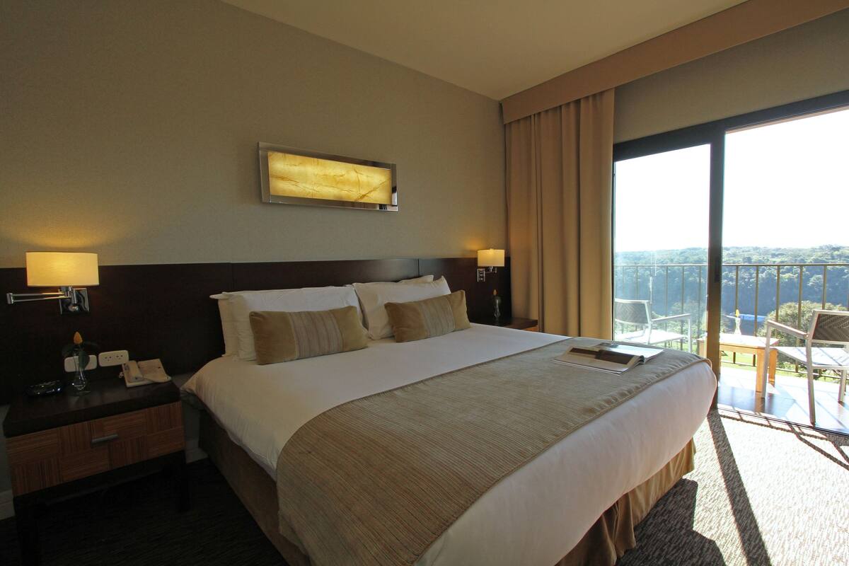 A hotel room with a large bed, bedside lamps, a nightstand, a view of the outdoors through a balcony.