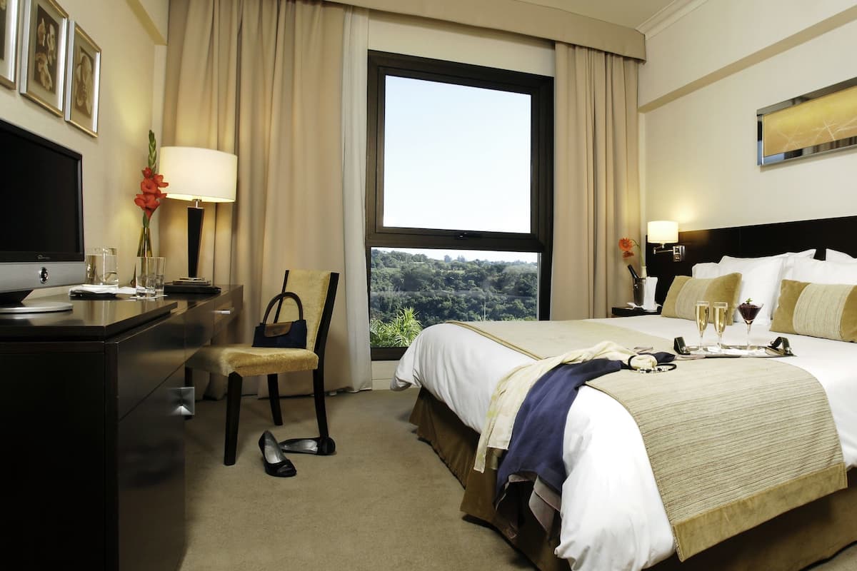 A hotel room with a bed, a desk, a chair, a TV, and a window with a view of trees.