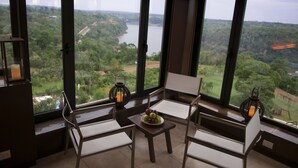 A room with large windows offering a view of a lake and forest. There are two chairs and a small table with a fruit bowl. - Panoramic Grand (Puerto Iguazú)