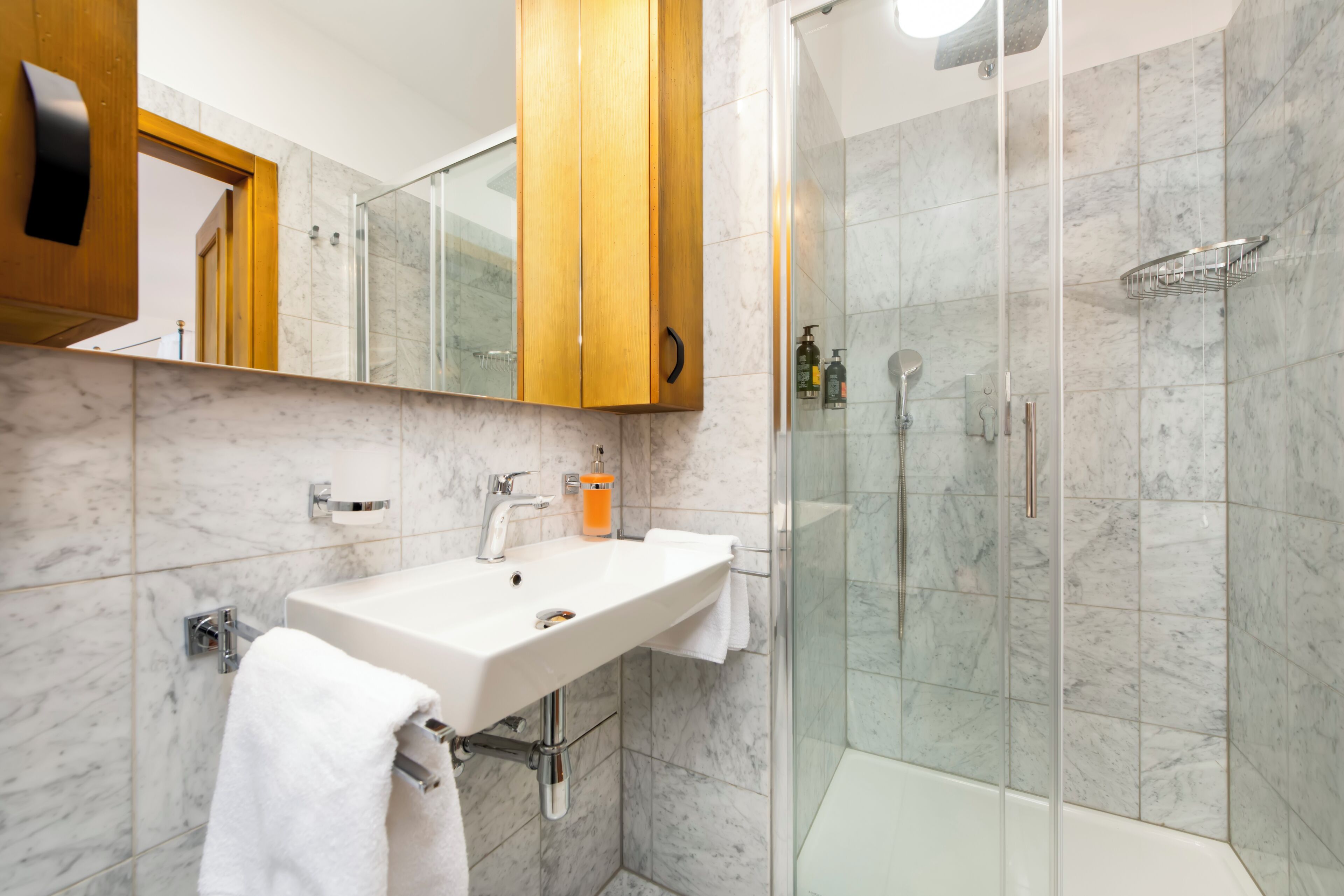 A modern bathroom with a marble-tiled shower, a white sink with a mirror above, and wooden cabinets.