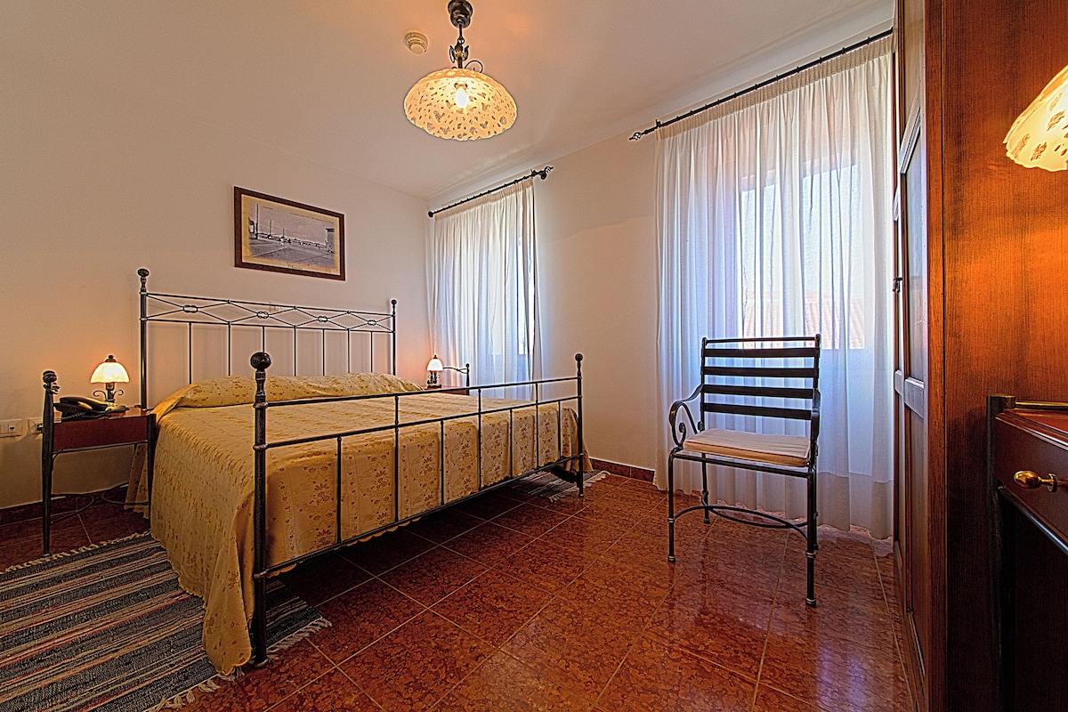 Un dormitorio con estructura de cama de metal, una silla, una mesita de noche con lámpara, un cuadro en la pared y una ventana con cortinas.
