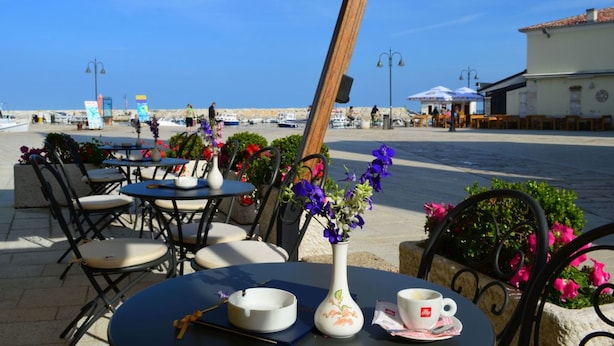 An outdoor café with tables and chairs, a vase with flowers, and a view of the sea and boats.