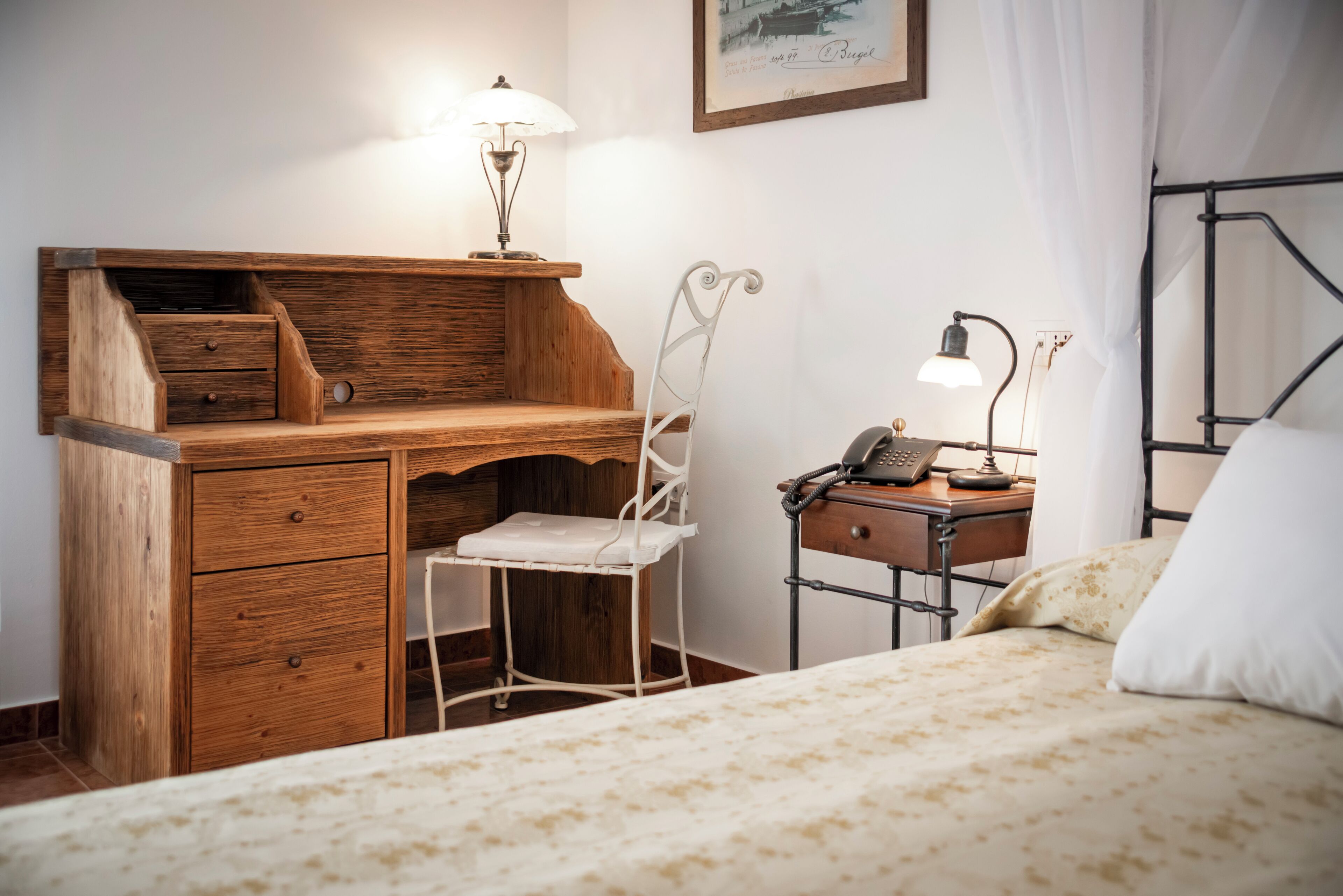 Ein Schlafzimmer mit einem Holztisch, einem weißen Stuhl, einem Bett mit gemustertem Überwurf, einer Nachttischlampe und einem gerahmten Bild an der Wand.