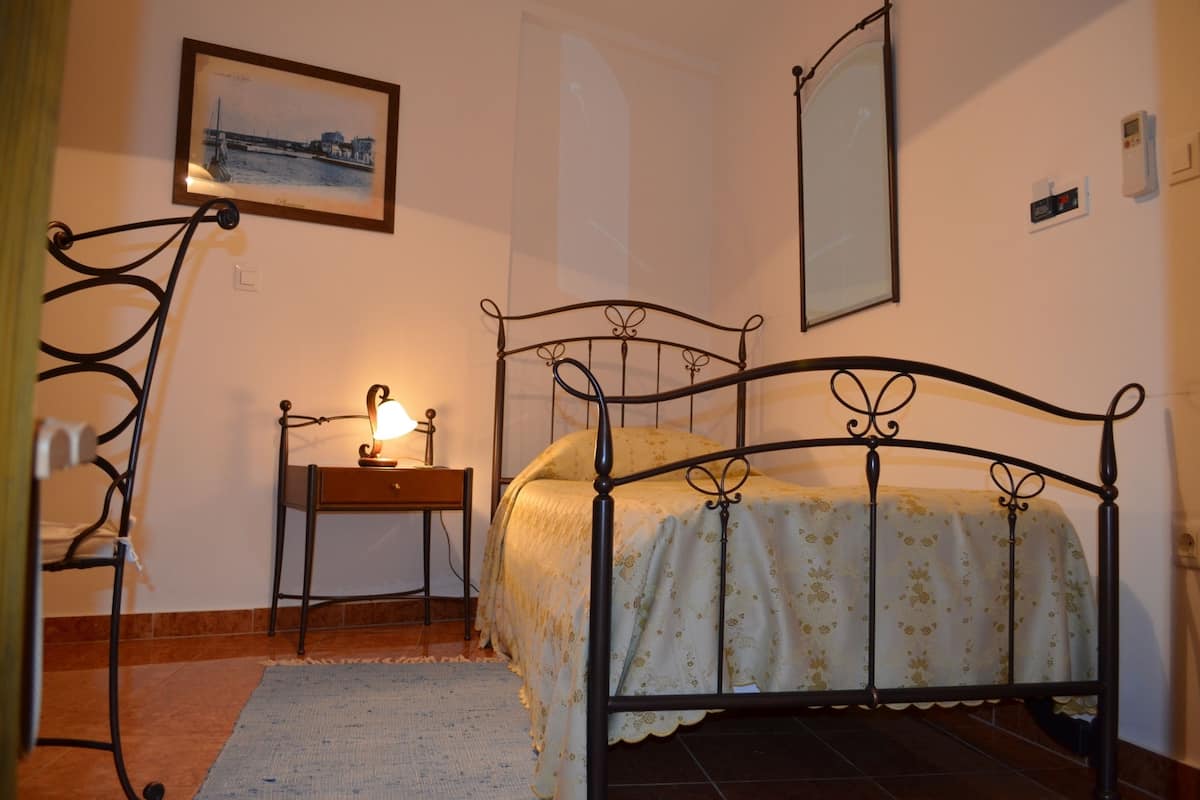 Een slaapkamer met een metalen bedframe, een nachtkastje met een lamp, een lijst met een foto aan de muur en een raam.