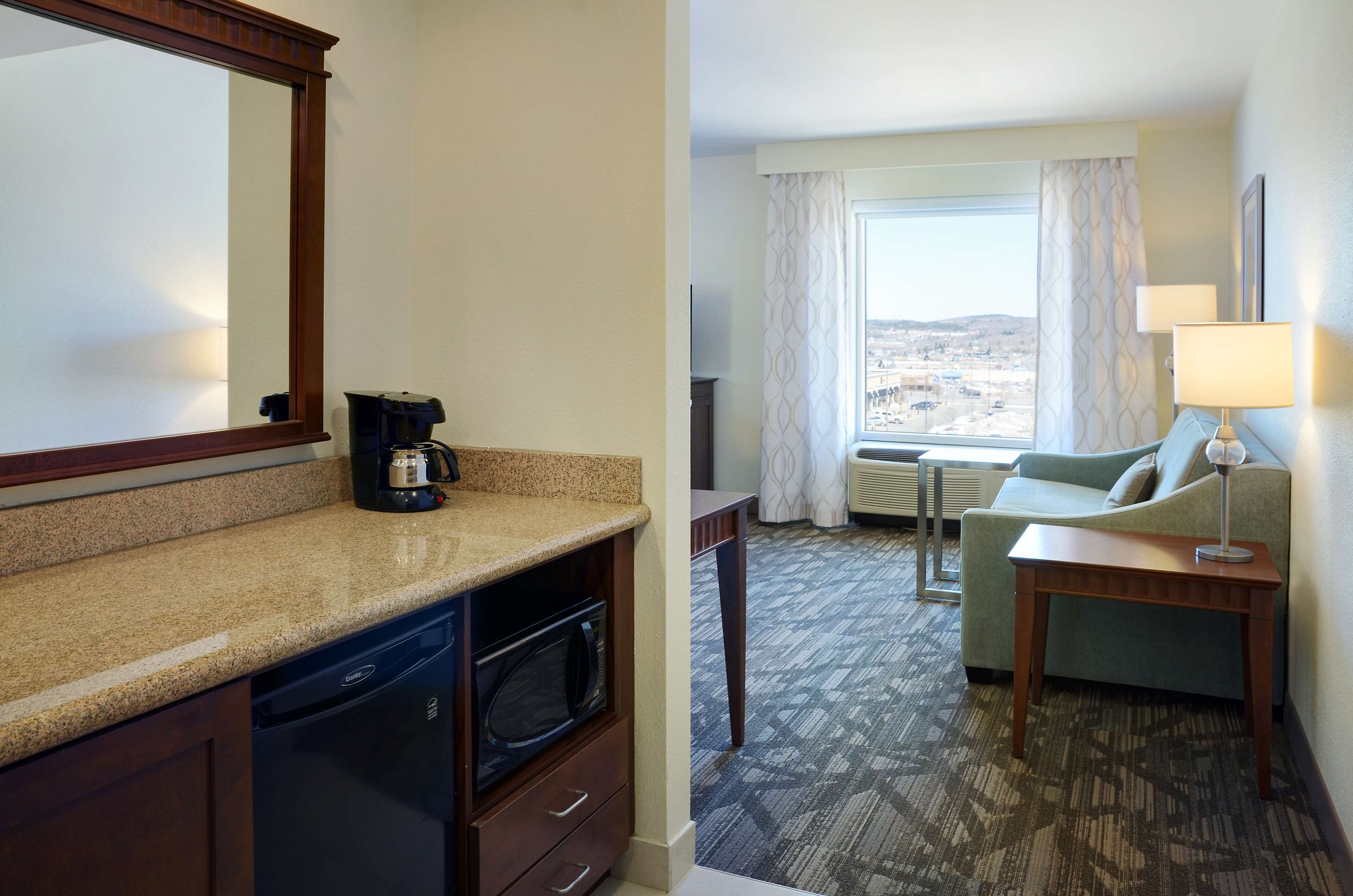 A hotel room with a kitchen area, a dining table, a sofa, and a view of the city through the window.