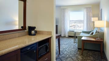 A hotel room with a kitchen area, a dining table, a sofa, and a view of the city through the window.