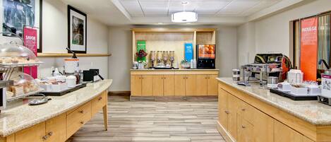 A clean, well-lit breakfast area with a counter, coffee machines, and a variety of food options.