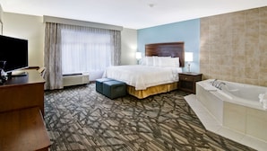 A hotel room with a large bed, a TV, a desk, a desk lamp, a bedside table, a lamp, a bathtub, and a window with curtains. - Hampton Inn & Suites Saint John (Saint John)