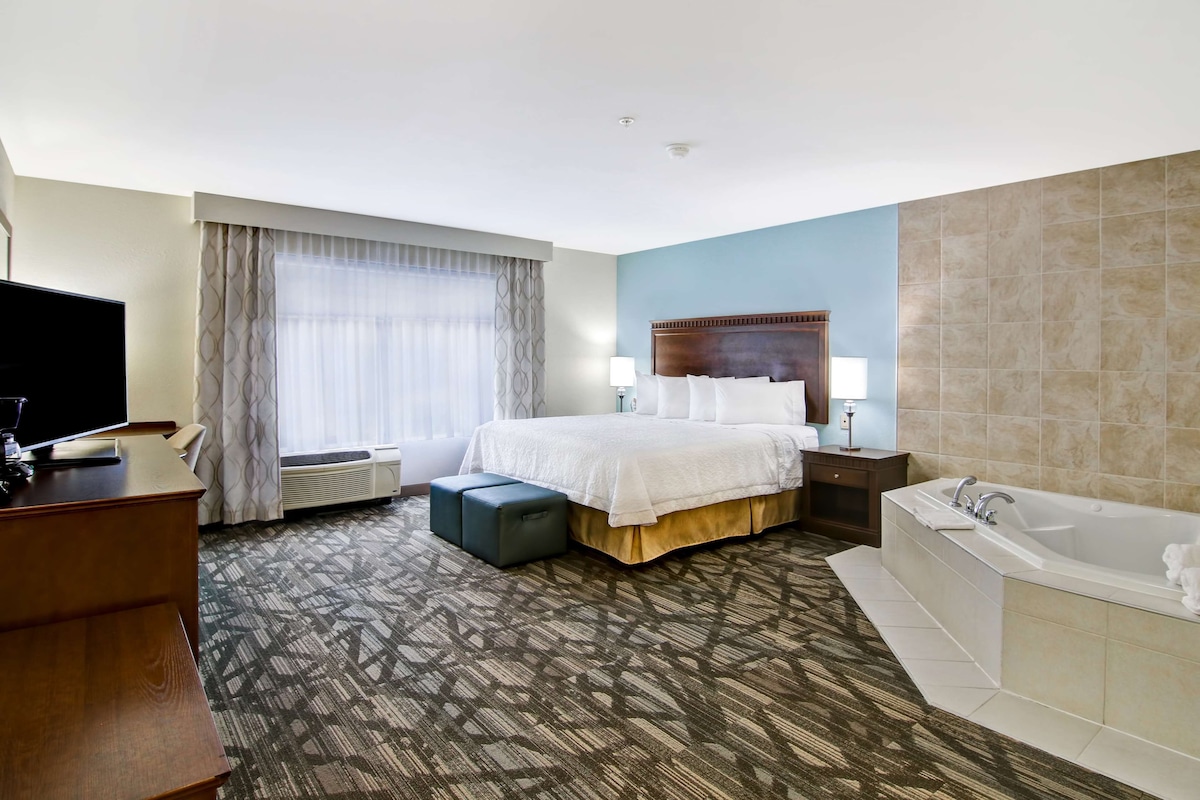 A hotel room with a large bed, a TV, a desk, a desk lamp, a bedside table, a lamp, a bathtub, and a window with curtains.