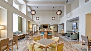 A spacious hotel lobby with a central dining area, sofas, and a kitchen in the background. - Hampton Inn & Suites Saint John (Saint John)