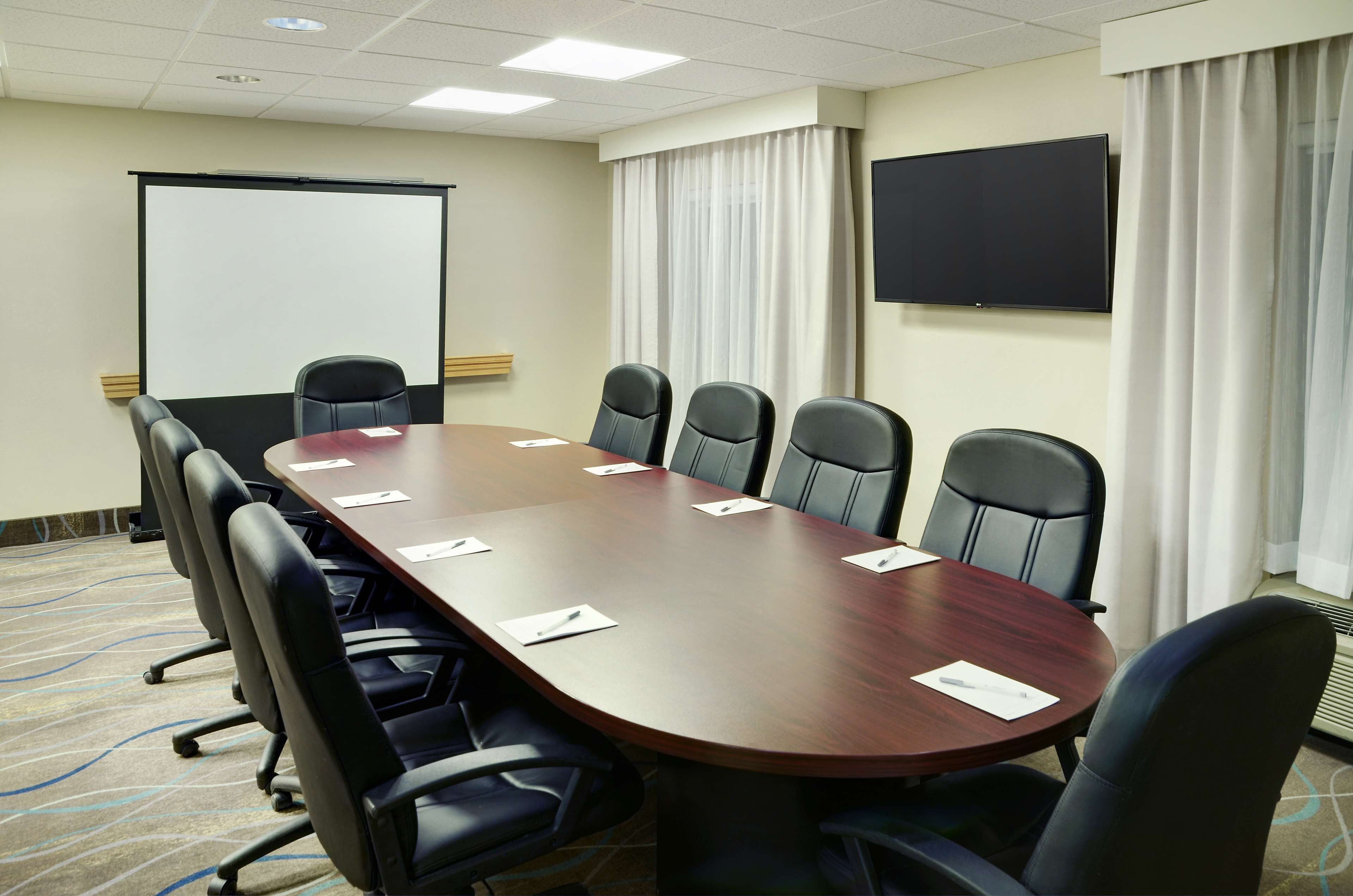 Un salón de conferencias con una larga mesa, sillas negras, una pantalla de proyector y un televisor.