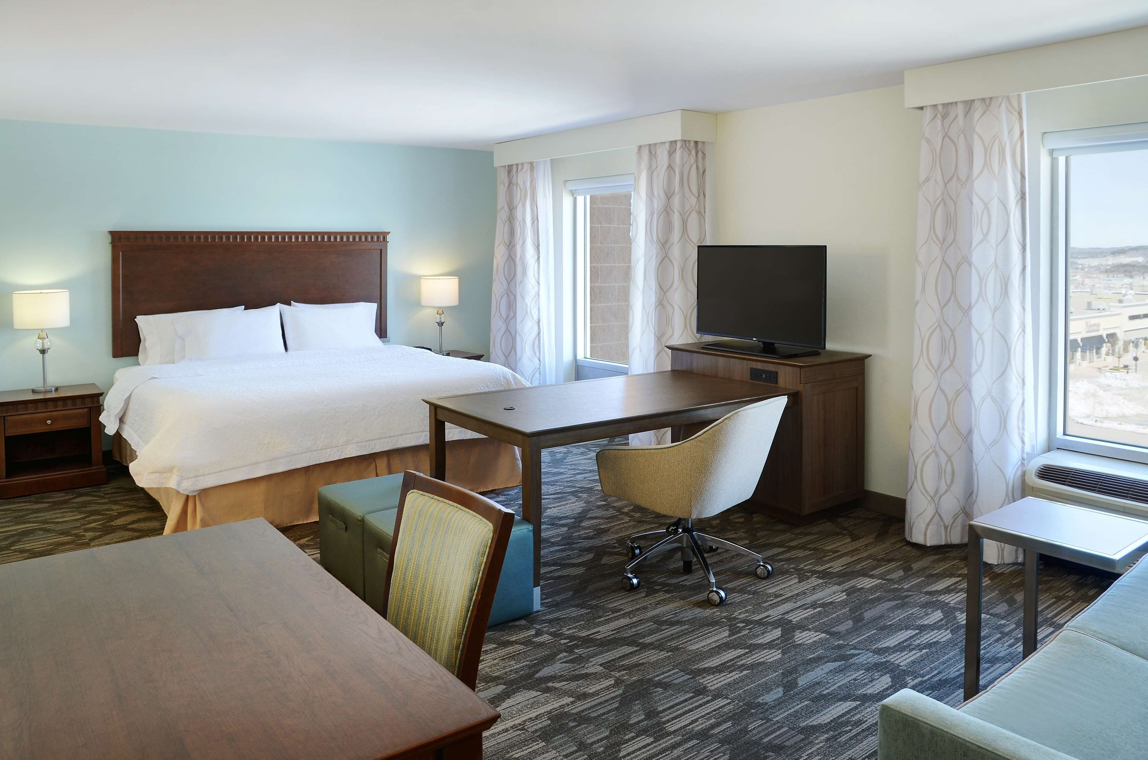 A hotel room with a large bed, a desk with a chair, a television, and a view of the city through the window.
