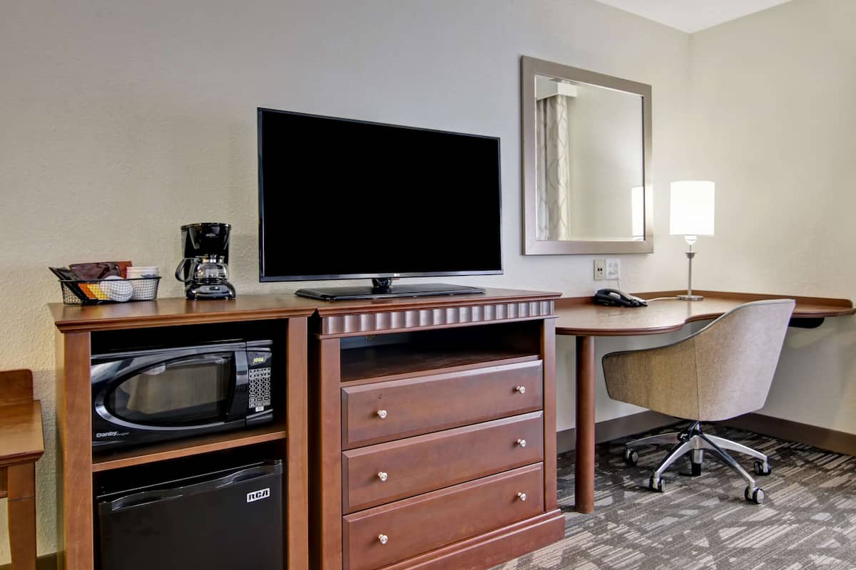 A hotel room with a flat-screen TV, a coffee maker, a microwave, a refrigerator, a desk with a chair, and a mirror.