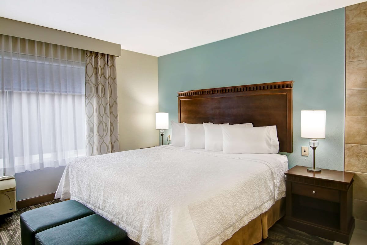 A hotel room with a large bed, two bedside tables with lamps, a bench, and a window with curtains.