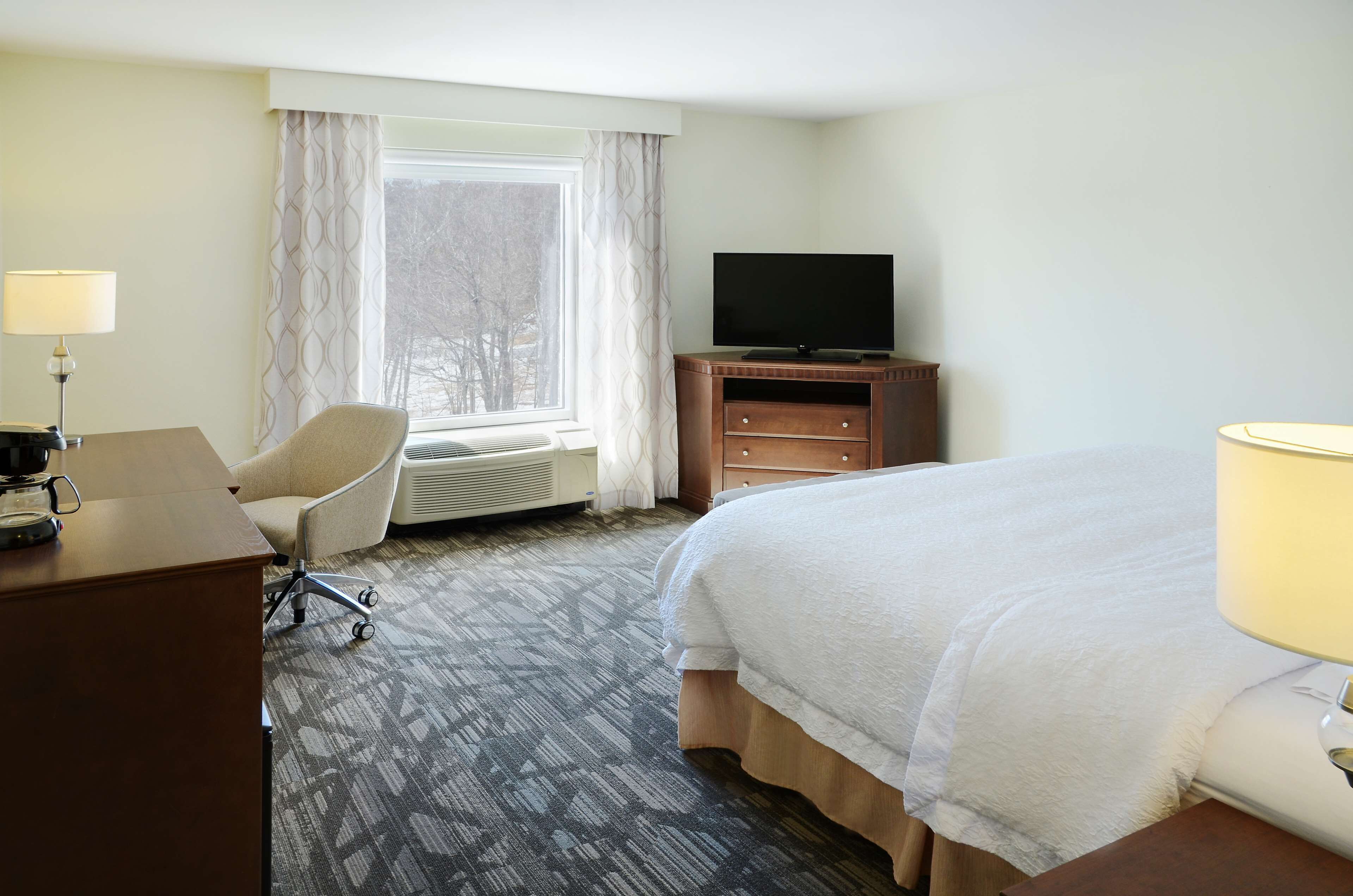 A hotel room with a bed, a desk with a lamp, a chair, a television on a wooden cabinet, and a window with curtains.