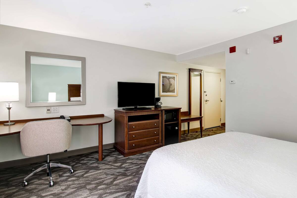 A hotel room with a large bed, a desk with a chair, a television, a dresser, and a mirror.