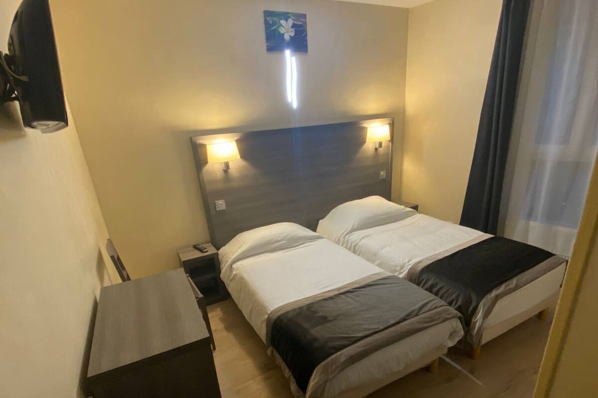 Ein Hotelzimmer mit zwei Betten, einem Schreibtisch, einem wandmontierten Fernseher und einem kleinen Fenster mit Vorhängen.