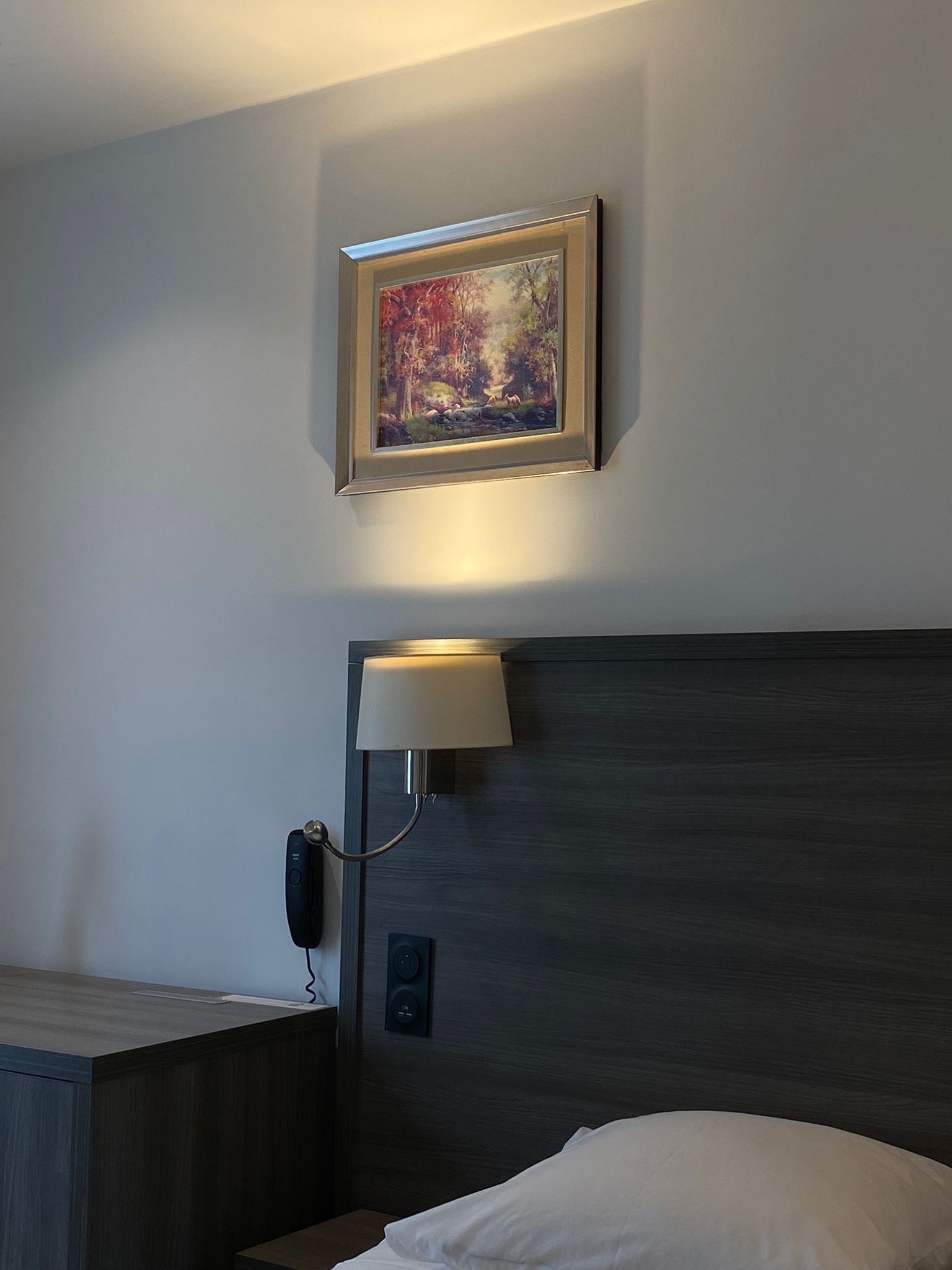 Habitación de hotel con cama, mesita de noche, lámpara, teléfono y un cuadro en la pared.