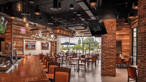 Un restaurante con una barra de madera alargada, varias mesas y sillas. Hay luces colgantes y un televisor montado en la pared.