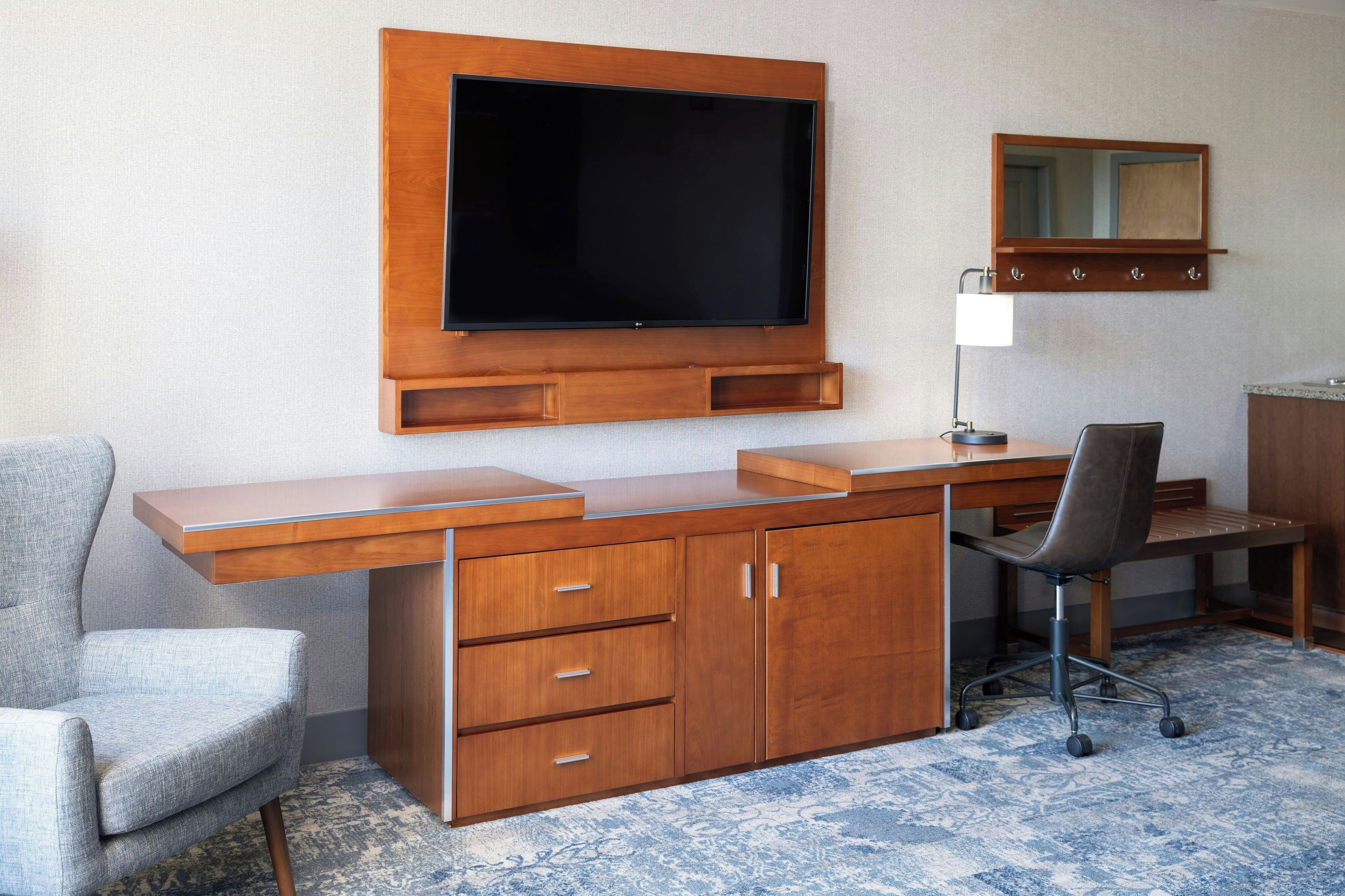 Une chambre d’hôtel avec un bureau en bois, un téléviseur à écran plat fixé au-dessus, une chaise, une lampe de bureau et un miroir au mur.