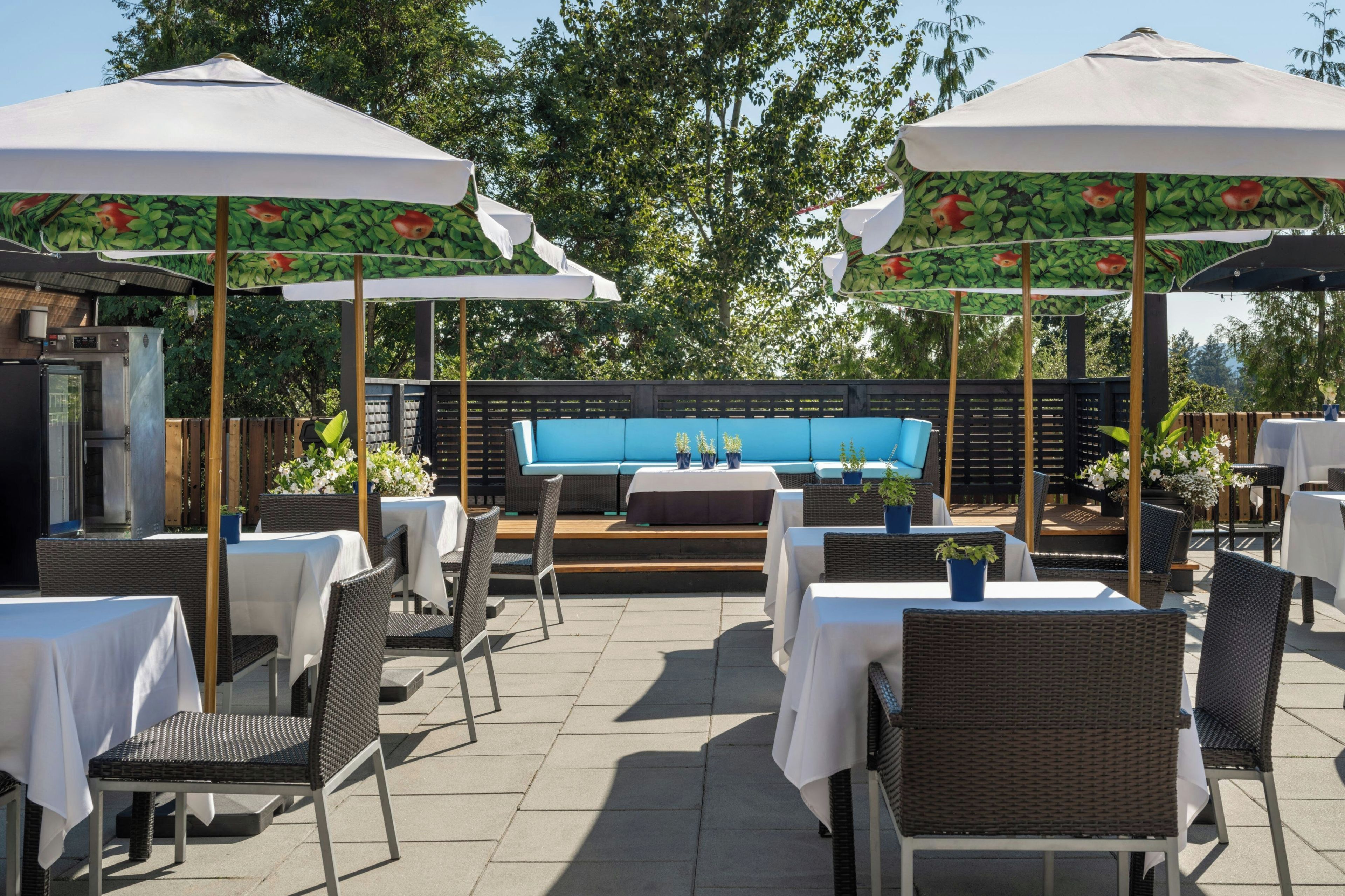 An outdoor patio area with tables and chairs, covered by umbrellas with fruit patterns, and a blue sofa in the background.