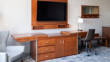 A hotel room with a wooden desk, a chair, a television mounted on the wall, a desk lamp, and a mirror above the desk.