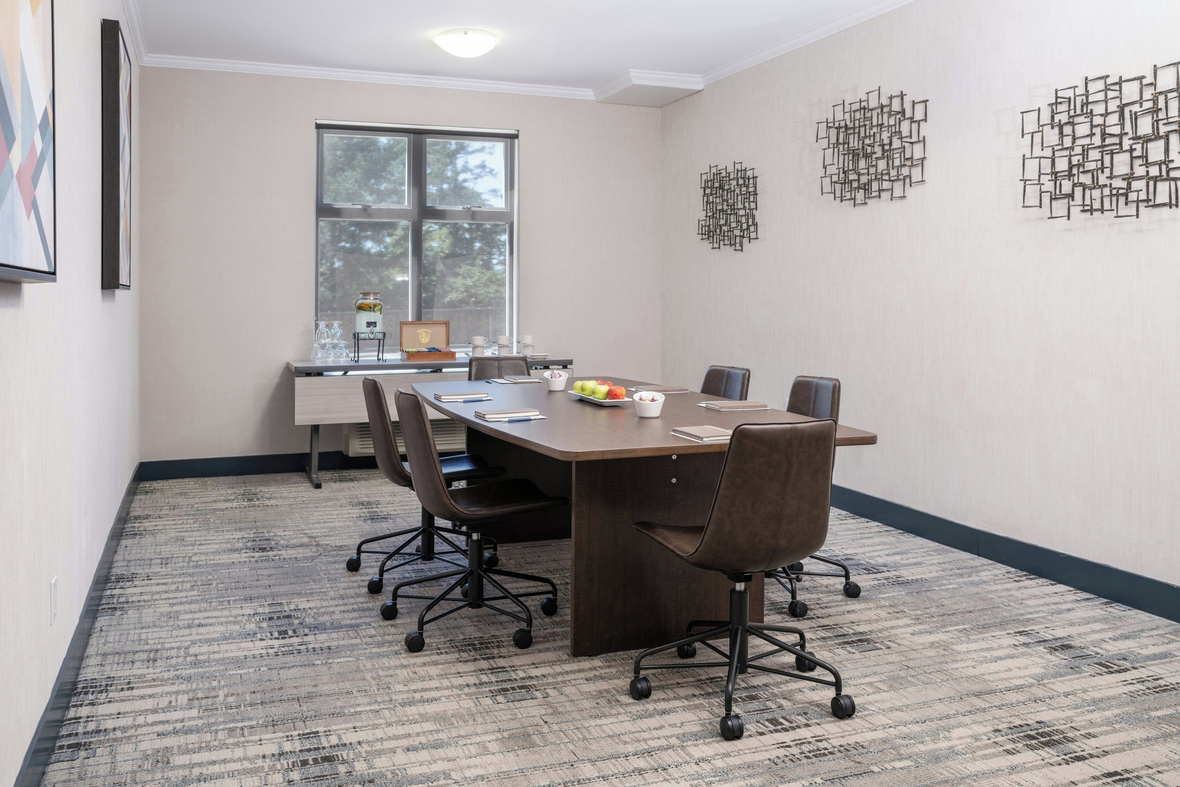 Un salón de conferencias con una mesa larga, seis sillas, un rincón para café y un ventanal con vistas a árboles.
