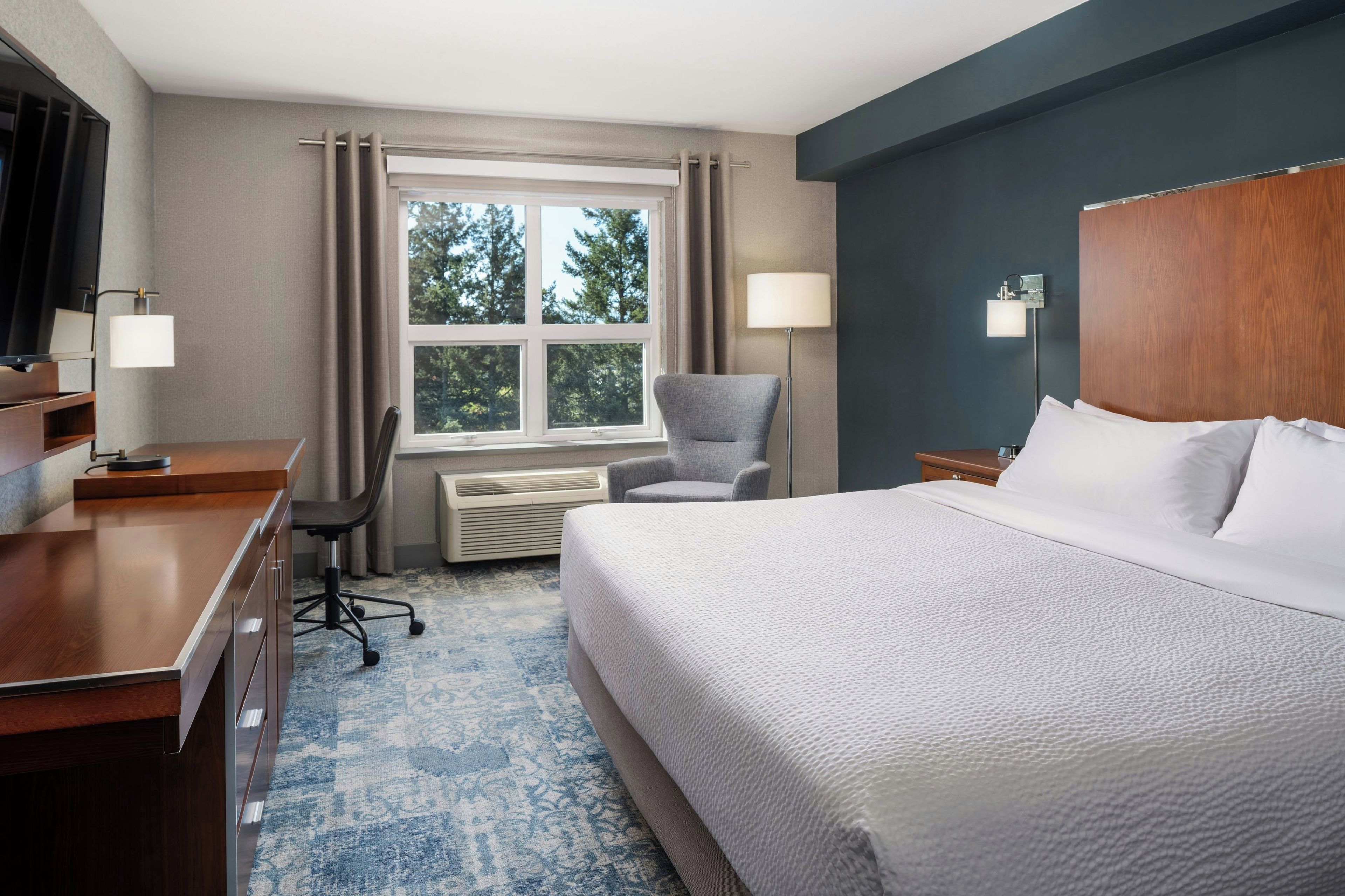 Quarto de hotel com cama, escrivaninha, cadeira e janela com cortinas.
