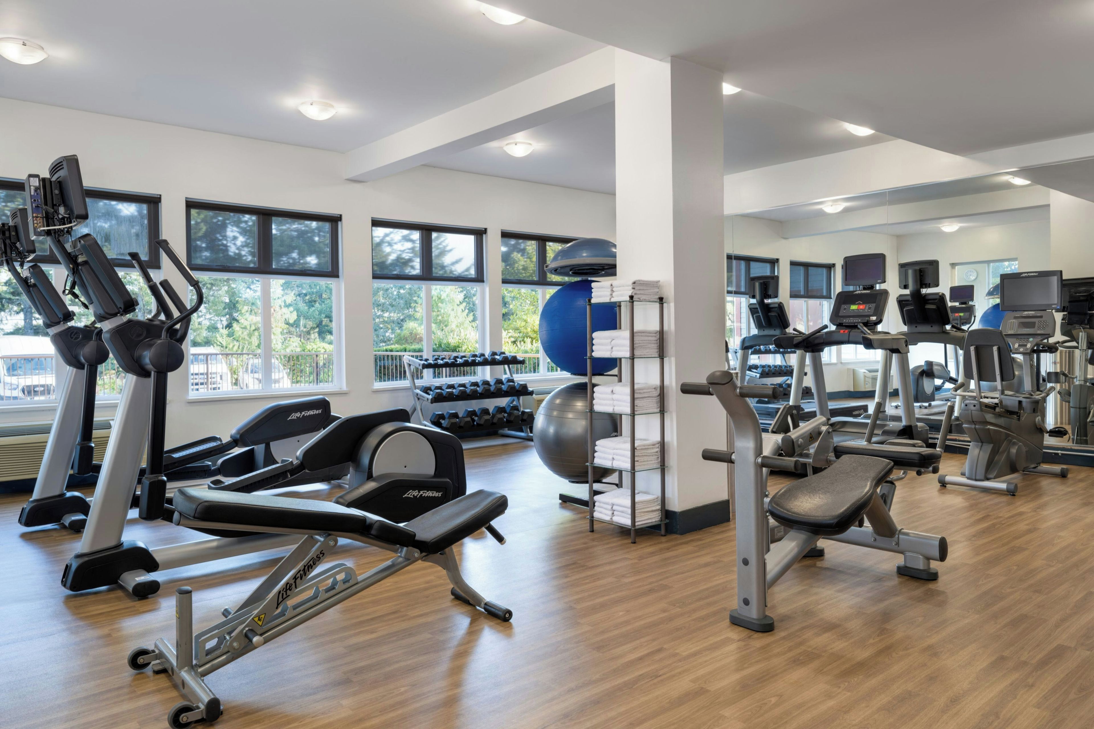 Un gimnasio con diverso equipamiento de ejercicio, amplias ventanas y suelo de madera.