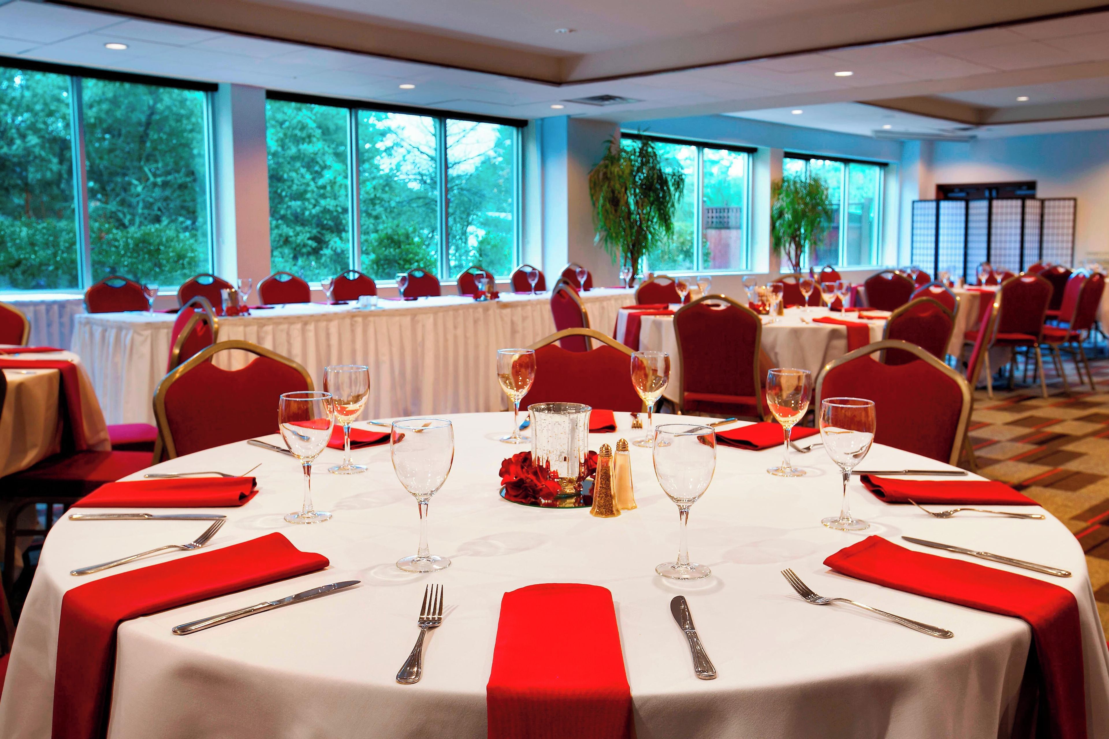 Uma sala de banquetes com mesas redondas preparadas para refeições, cada uma com toalhas brancas, guardanapos vermelhos e talheres.