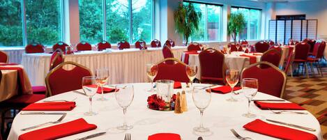 Uma sala de banquetes com mesas redondas preparadas para refeições, cada uma com toalhas brancas, guardanapos vermelhos e talheres.