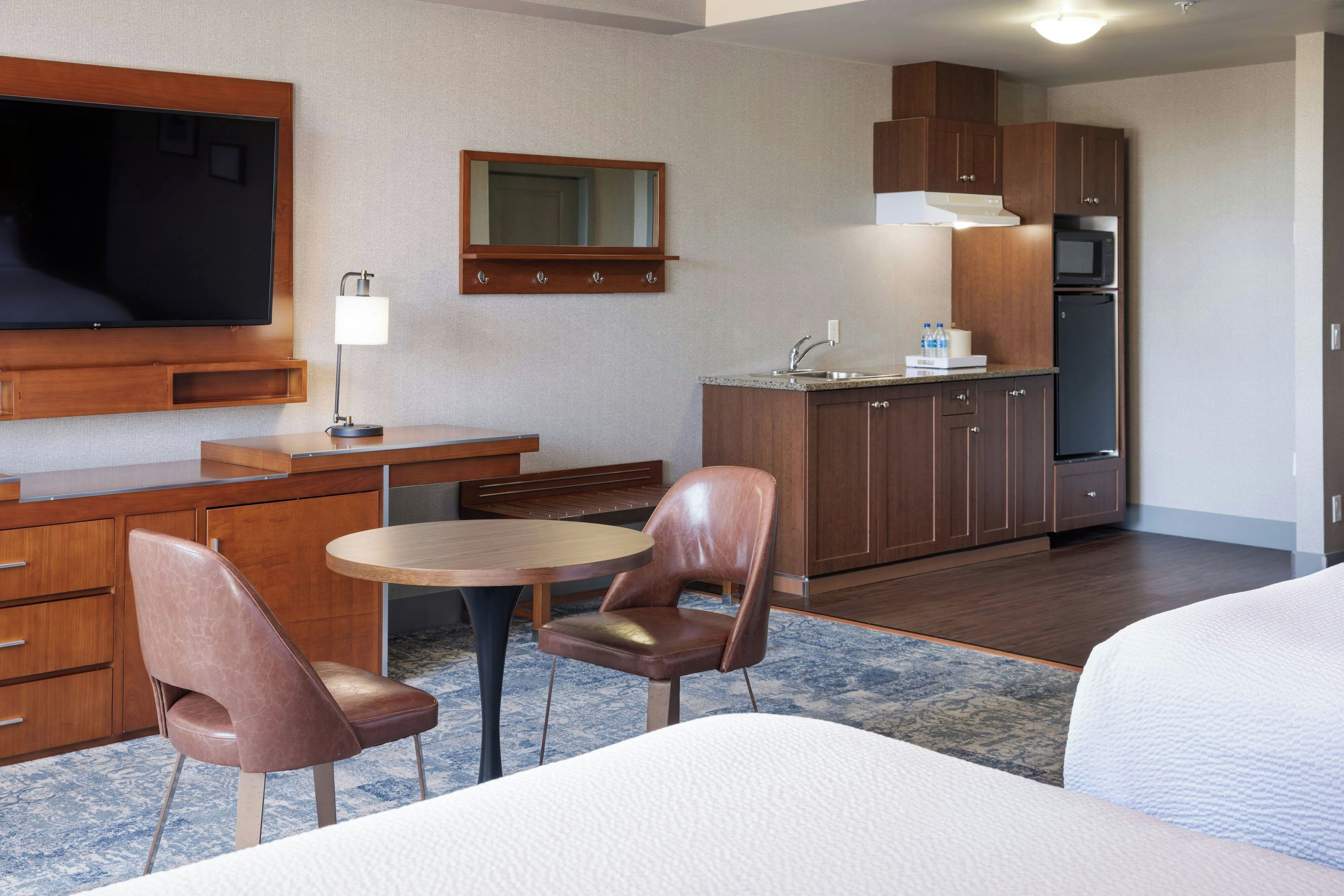 A hotel room with a bed, a desk with a chair, a television, a mirror, a kitchenette, and a bathroom.