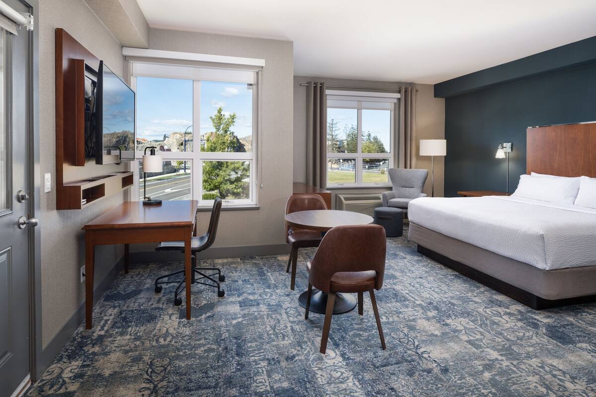 A hotel room with a bed, a desk, two chairs, a TV, and large windows offering a view of the outdoors.