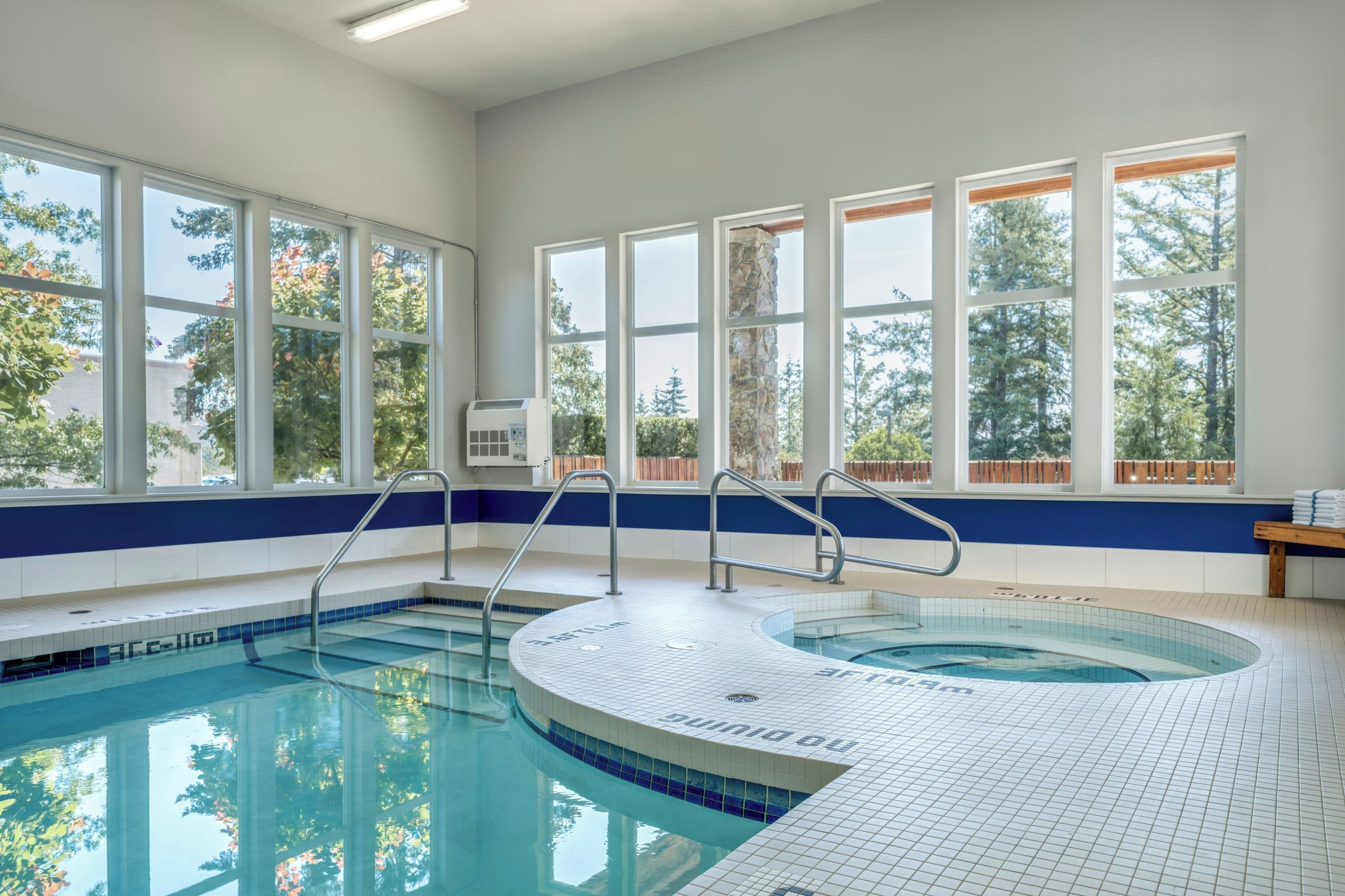 An indoor swimming pool with clear blue water, surrounded by large windows offering views of trees and a building.