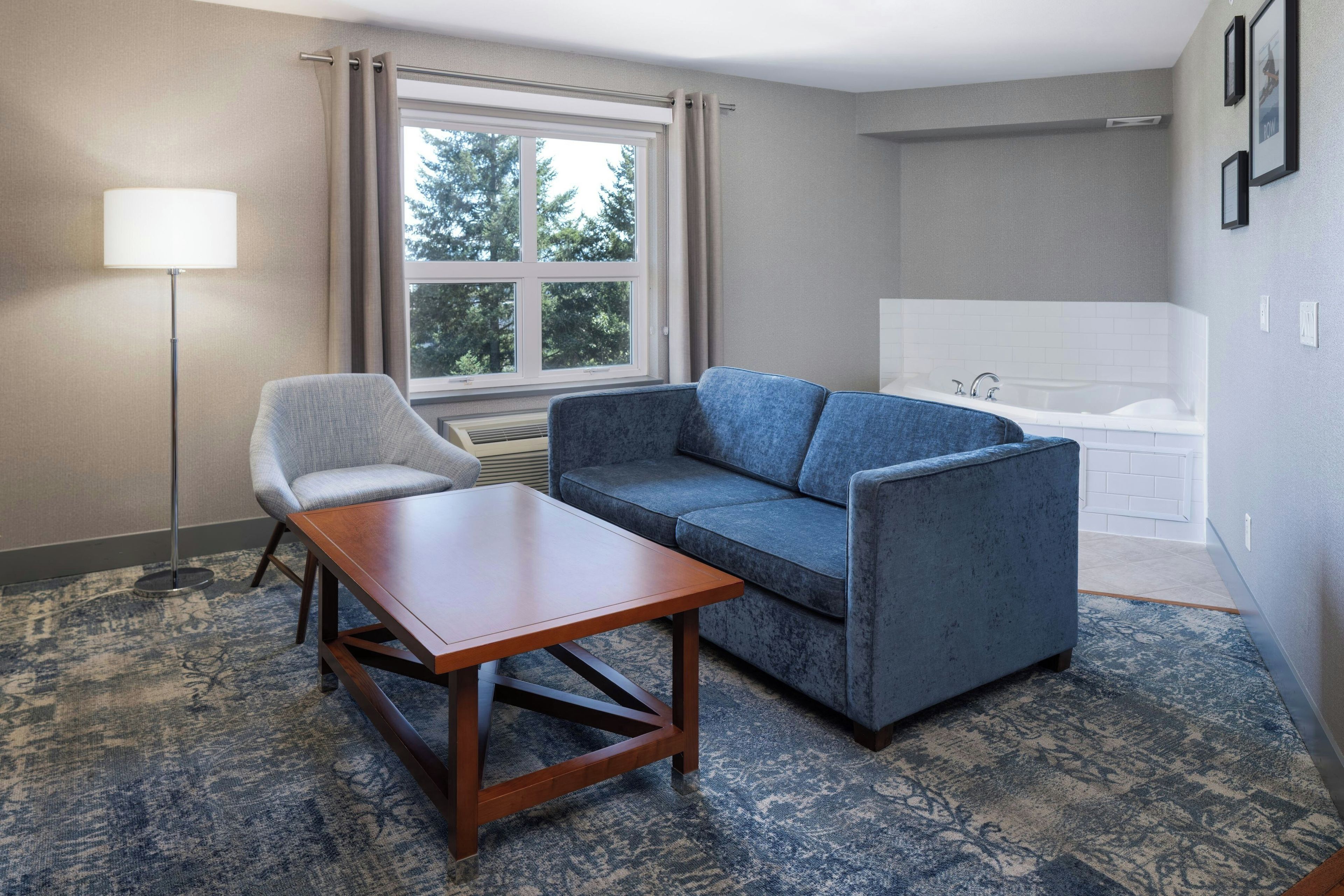 A hotel room with a sofa, a small table, a lamp, a window with curtains, and a bathroom with a bathtub.