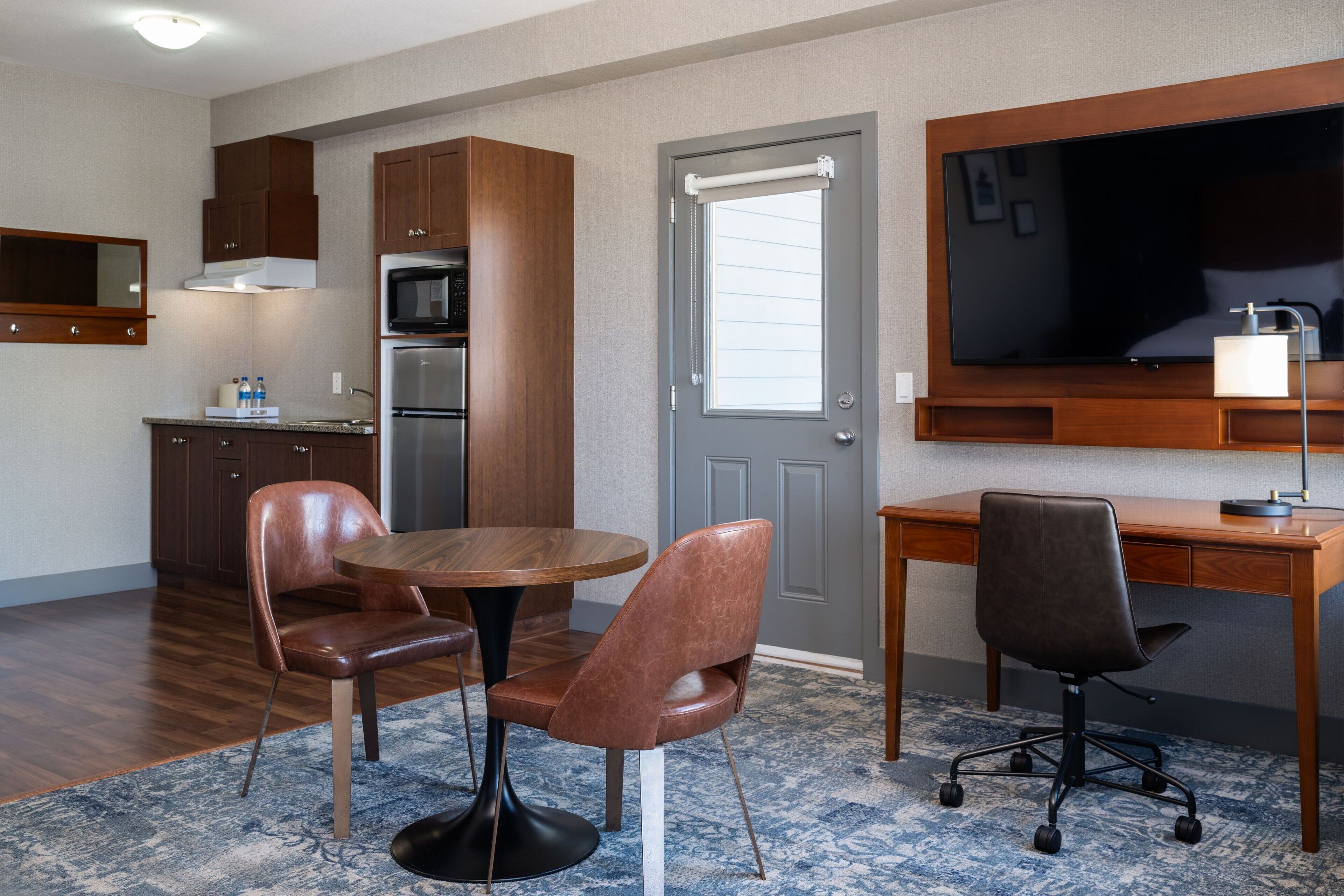 Une chambre d’hôtel compacte comprenant un coin cuisine, un espace repas avec deux chaises, un bureau avec une chaise et un grand téléviseur à écran plat fixé au-dessus du bureau.