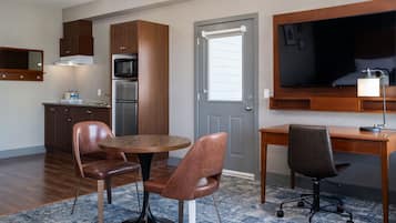 Habitación de hotel compacta con kitchenette, zona de comedor con dos sillas, escritorio con silla y un televisor de pantalla plana grande montado sobre el escritorio.