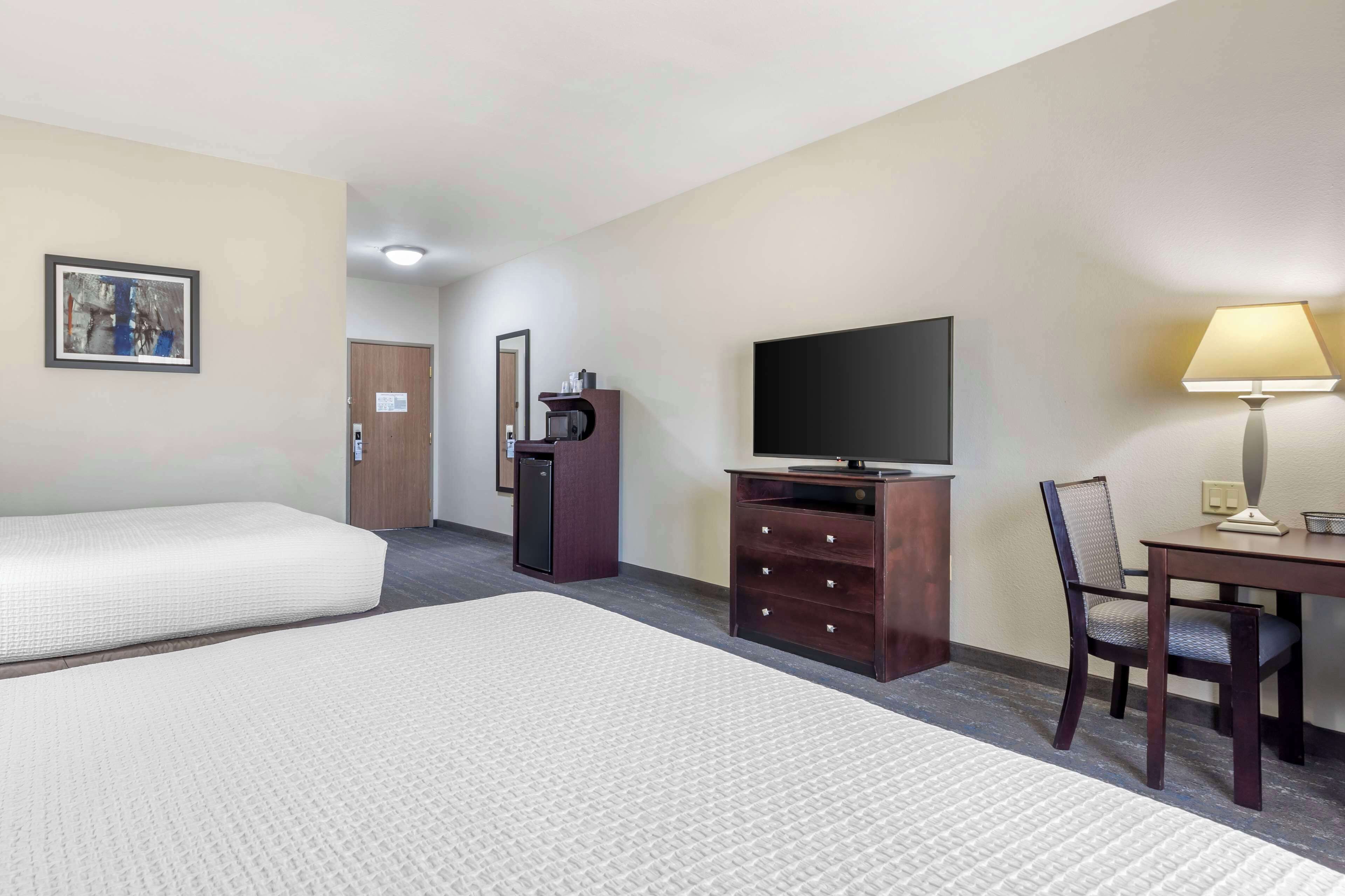A hotel room with two beds, a desk, a TV, a lamp, a small refrigerator, and a door in the background.