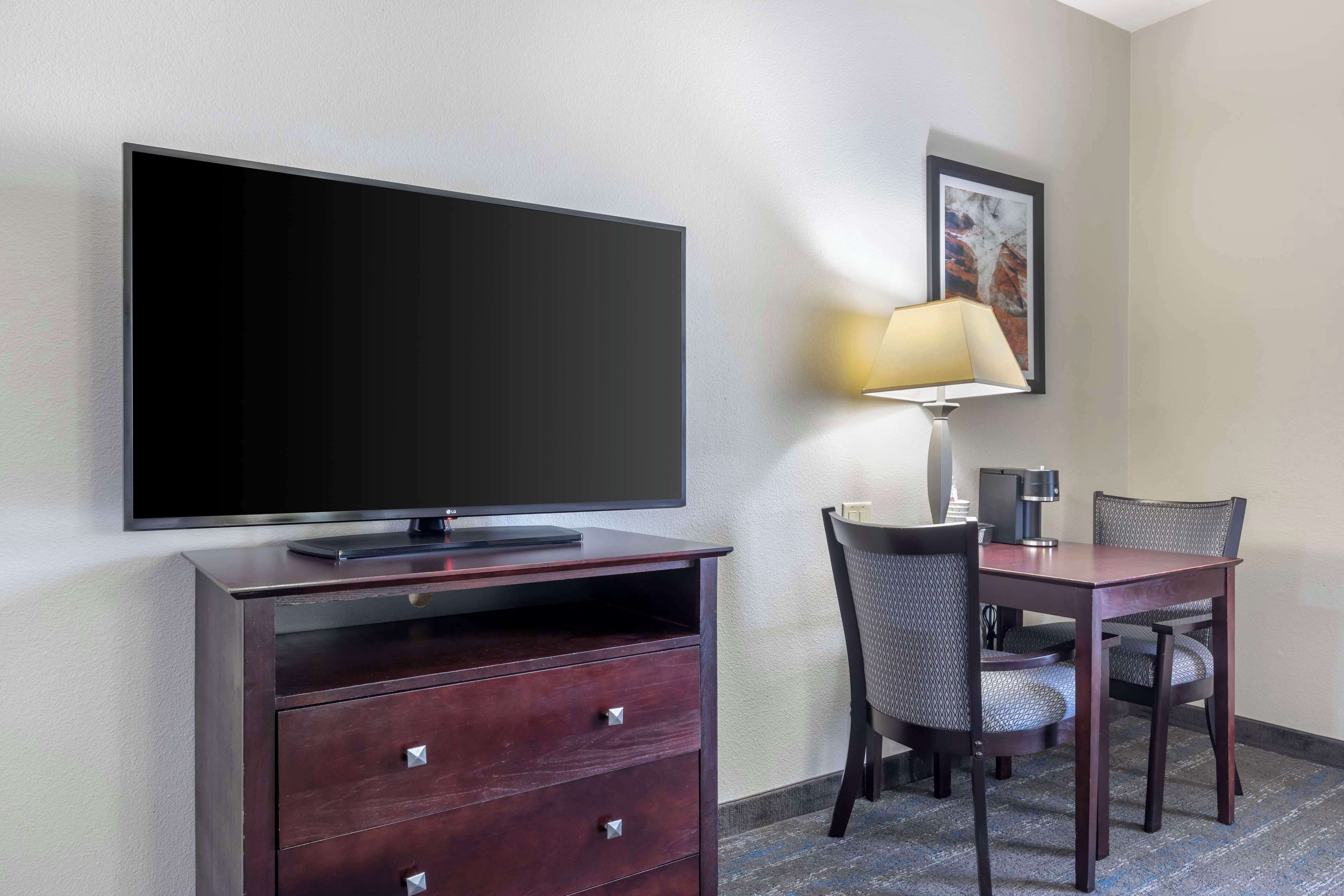 Habitación de hotel con televisor de pantalla plana, cómoda de madera, mesa de comedor con dos sillas, lámpara y un cuadro en la pared.
